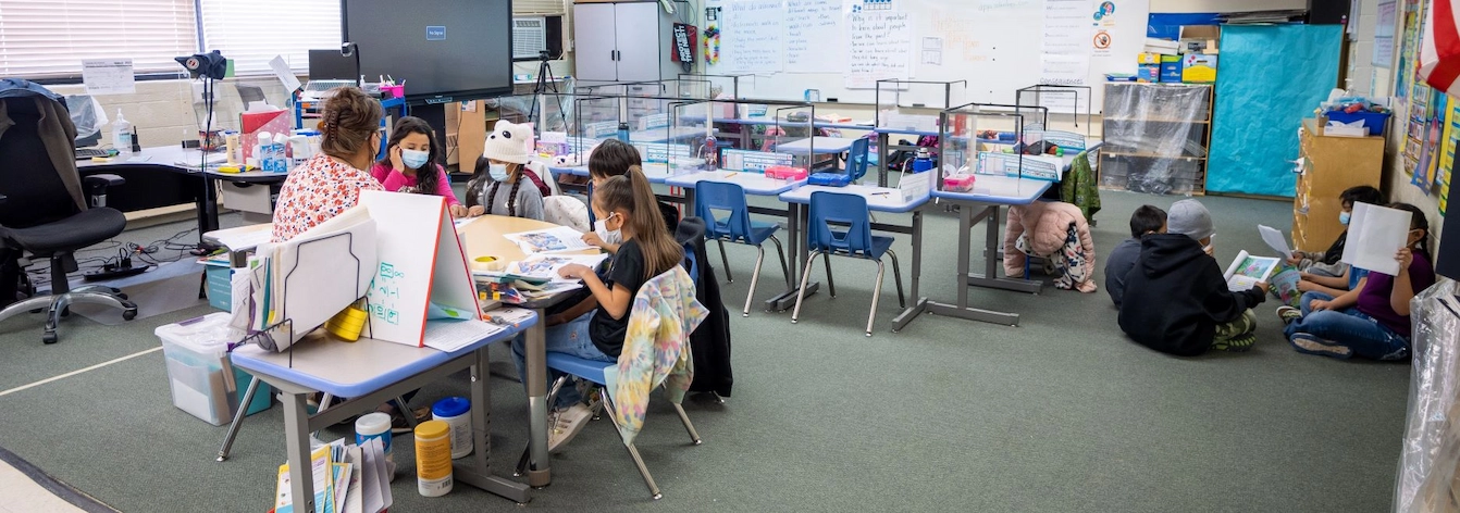 Group of students and teacher at table in classroom and another group of students reading on floor