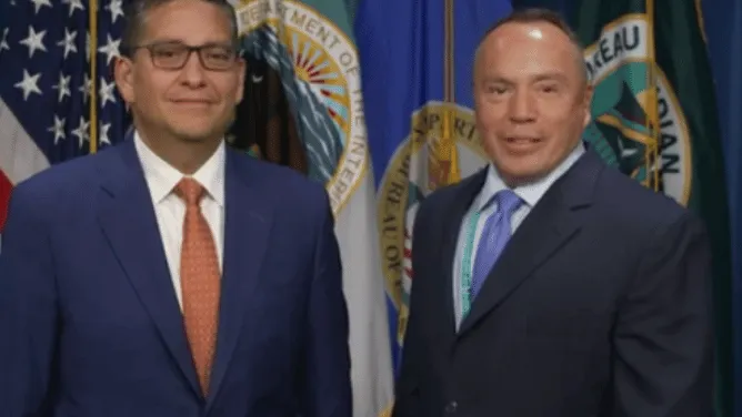 Billy Kirkland and Director Dearman stand side by side in front of U.S. government and BIE flags.